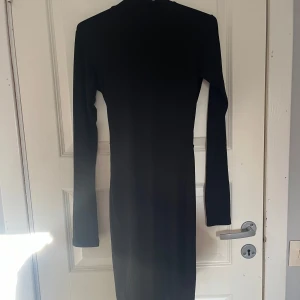 Svart långärmad klänning med öppen rygg - Svart ribbad långärmad klänning med halvpolo och snygg öppen rygg och knytband i ryggen. Klänningen är figurnära och har en längre passform till knäna ungefär. Sparsamt använd, hör av er vid frågor