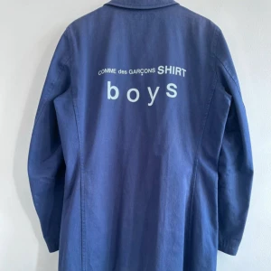 Blå rock från Comme des Garçons - Snygg blå rock från Comme des Garçons SHIRT boys i 100% bomull. Jackan har fyra stora fickor framtill, klassisk krage och knäppning med tre knappar. Tryck med logga på ryggen ger en cool vibe. Perfekt för lager-på-lager och streetstyle. Nypris 7000kr, pris kan diskuteras.