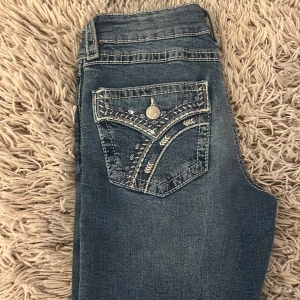 Blå jeans med broderad bakficka - Snygga blå jeans med en unik broderad bakficka och dekorativa sömmar. Fickan har även en liten metallknapp och detaljer i vitt och blått. Perfekta för dig som vill sticka ut med coola detaljer.