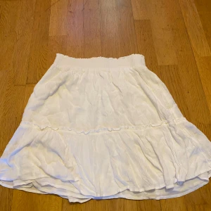 Vit volangkjol L Vera Skirt S - Supersöt vit kortkjol från L Vera med volang och resår i midjan. Kjolen har en lätt rynkad struktur och är perfekt för dig som gillar en luftig och feminin stil. Passar perfekt till sneakers eller sandaler.