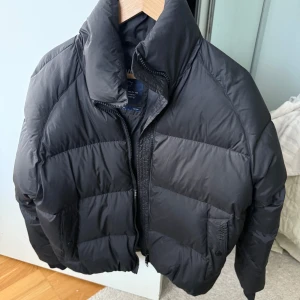 Svart pufferjacka från Svea, M - Säljer en svart pufferjacka från Svea i storlek medium. Jackan har hög krage, dragkedja framtill och är riktigt voluminös. Yttertyget är i polyester och fodret i nylon, med vaddering av dun och fjäder. Perfekt för kalla dagar när du vill hålla värmen och ändå se snygg ut.