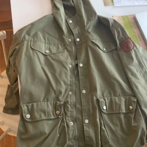 Grön vindjacka från Fjällräven - Säljer en grön vindjacka från Fjällräven med huva, stora fickor med tryckknappar och klassisk logga på ärmen. Jackan har dragkedja och knappar framtill, samt en stilren och funktionell design perfekt för uteliv.