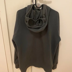 Mörkgrå hoodie med goggles från C.P. Company - Säljer en mörkgrön zip hoodie från C.P. Company med dragkedja och goggles i huvan. Tröjan är i ett väldigt bra skick. Sitter som en M. Hör av dig vid frågor! 