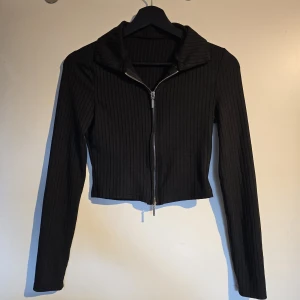 Svart ribbad croppad kofta - Säljer en svart ribbad kofta med croppad passform och lång ärm. Tröjan har en snygg krage och dragkedja framtill som gör den enkel att styla. 