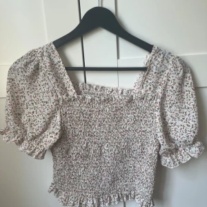 Blommig puffärm blus från SHEIN - Supersöt blommig blus från SHEIN med puffärmar och smockad kropp. Toppen är vit med små rosa och gröna blommor, har volangdetaljer vid ärmslut och nederkant samt fyrkantig ringning. Perfekt för dig som gillar romantisk och trendig stil.