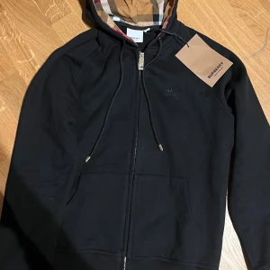 Svart hoodie från Burberry med dragkedja - Hej! Säljer en svart hoodie från Burberry med klassiskt rutigt foder i huvan, dragkedja framtill och snörning vid huvan.  Broderad Burberry-logga på bröstet.   Tillverkad i mjuk bomull för en skön känsla. Perfekt för dig som vill ha en stilren och lyxig look.  Obs!  Storlek M men passar snarare som S!!  Vill du veta något mer eller vill ha fler bilder så är det bara att skriva! Priset är såklart inte hugget i sten