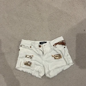 Vita paljettshorts från DKNY - Snygga vita shorts från DKNY med råa kanter och guldiga paljett-detaljer framtill. Klassisk femficksmodell i jeansmaterial med broderad DKNY-logga bak och metallicmärke i midjan. Perfekta för sommarens alla äventyr.