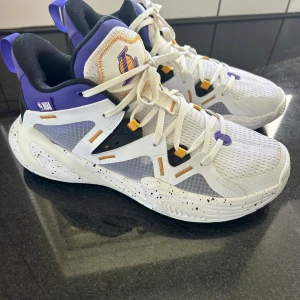 Los Angeles Lakers sneakers NBA - Vita NBA skor med lila och gula detaljer. Sulan är vit med svarta stänk. Skorna har mesh-ovandel, snörning och Lakers-logga på plösen. Perfekt för basket eller fritid.  Lappen är bortklippt men innermåttet är 27 cm.