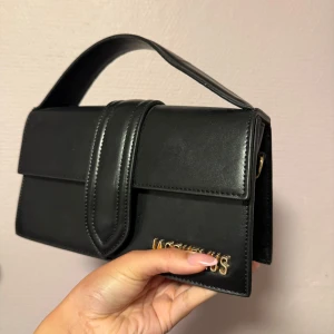 Svart handväska från JAQ i skinn - Stilren svart handväska från J med guldiga bokstäver på framsidan. Väskan har en rektangulär form, bredt handtag och magnetlås. Utsidan är i slätt skinn och insidan har en mjuk beige yta. Perfekt för dig som gillar klassisk design med en lyxig touch.