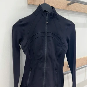 Lululemon define jacket nulu - Snygg svart träningsjacka från Lululemon med figurnära passform och hög krage. Jackan har hel dragkedja framtill, två fickor med dragkedja och långa ärmar. Perfekt för gymmet eller powerwalks. Stilren design med diskret logga på dragkedjan. Storlek 2 motsvarar xs slutsåld 