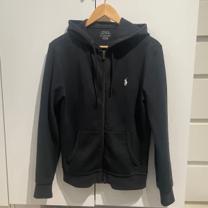 Svart hoodie från Polo Ralph Lauren - Svart hoodie från Polo Ralph Lauren med dragkedja framtill, huva med snörning och klassisk vit broderad logga på bröstet. Mjuk bomullskänsla och två fickor framtill. Perfekt för en chill och stilren look.