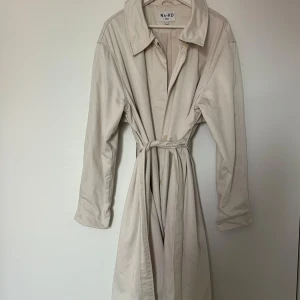 Beige trenchcoat från NA-KD - Stilren beige trenchcoat från NA-KD med klassisk krage, knäppning framtill och avtagbart bälte i midjan. Jackan har långa ärmar och två fickor på sidorna. Perfekt för lager-på-lager och en clean look.