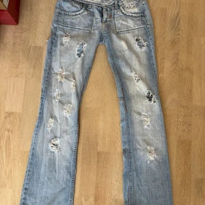 Ljusblå slitna jeans med tryck - Säljer ett par ljusblå jeans med rejält slitna detaljer och hål på både fram- och baksida. Jeansen har bootcut-modell och ett orange tryck med blommor på ena bakfickan. Klassisk femficksdesign och cool vintagekänsla. Storlek 34/36!🩷