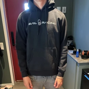 Svart hoodie från Sail Racing - Svart hoodie från Sail Racing med vit logga på bröstet och en mindre logga på huvan. Tröjan har snörning i huvan, stor magficka och ribbade muddar vid ärmslut och nederkant. Perfekt för chill dagar och snygg till jeans.