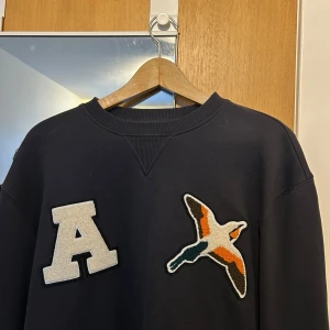 Mörkblå sweatshirt från Axel Arigato med fågelpatch - Mörkblå sweatshirt från Axel Arigato i 100% bomull. Framsidan har en stor vit A-patch och en broderad fågel i orange, grön och vit. Tröjan har rund hals och långa ärmar, perfekt för dig som gillar streetwear med coola detaljer.