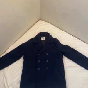 Mörkblå skepparkavaj från C Studio - Stilren mörkblå skepparkavaj från C Studio med dubbelknäppning och klassisk krage. Jackan har två snedställda fickor framtill och är tillverkad i ullblandning som håller dig varm under kyliga dagar. Perfekt för dig som gillar en tidlös och clean look.