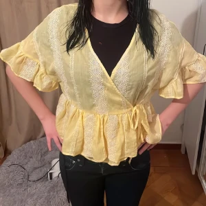 Zara Gul omlottblus med volangärm - Söt gul Zara omlottblus med knyt i sidan och volangärmar. Blusen har broderade spetsdetaljer och ett luftigt, lätt material som passar perfekt till vår och sommar. Den har korta ärmar och en avslappnad passform med markerad midja. Tryck köp nu