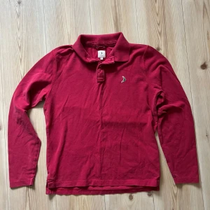 Röd långärmad pikétröja Boomerang M - Röd långärmad pikétröja från Boomerang med klassisk krage och knappslå framtill. Tröjan har en liten broderad logga på bröstet och är tillverkad i mjuk bomull. Perfekt för en chill och stilren look.