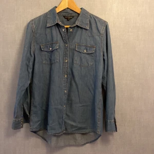 Jeansskjorta från KappAhl, stl 42 - Snygg jeansskjorta från KappAhl i klassisk blå denim. Skjortan har två bröstfickor med lock och tryckknappar, långa ärmar med knappar vid manschetten och en rak passform. Perfekt för en avslappnad och cool look.