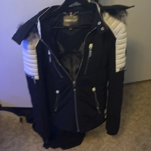 Svart vinterjacka med päls från Horspist - Snygg svart vinterjacka från Horspist med vita quiltade partier på axlar och ärmar. Jackan har huva med fluffig fuskpäls, flera dragkedjor och coola detaljer. Perfekt för kalla dagar och ger en riktigt urban vibe.