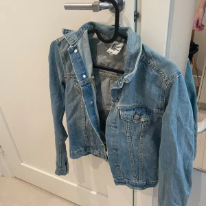 Klassisk blå jeansjacka H&M - Snygg blå jeansjacka från H&M med klassisk passform. Jackan har knappar framtill, två bröstfickor med lock och knapp samt långa ärmar. Perfekt att slänga över en hoodie eller t-shirt för en chill look.