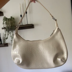 Beige axelväska i crocopräglat skinn - Snygg cremevit axelväska från Gina Tricot med crocopräglat mönster och dragkedja upptill. Väskan har en stilren, rundad form och ett kort axelband med silvriga metalldetaljer. Knappt använd
