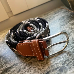 Flätat svart och grått bälte - Snyggt flätat bälte i svart, grönt  och vitt med detaljer i brunt konstläder och silvrigt spänne. Bältet har en cool, randig look och passar perfekt till jeans eller chinos. Flexibelt material som gör det enkelt att justera efter midjan.