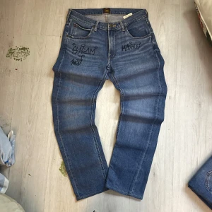 Blå Lee jeans med graffiti-detaljer - Säljer ett par klassiska blå jeans från Lee med unika graffiti-liknande svarta tags på fram- och baksidan. Jeansen har rak passform, fem fickor och Lee-läderpatch bak i midjan. Perfekta för dig som vill sticka ut med en personlig touch.