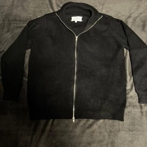 Svart ribbad kofta Maison Margiela - Svart ribbstickad kofta från Maison Margiela med hel dragkedja framtill och klassisk krage. Tröjan är långärmad och har en clean, minimalistisk look med diskreta signaturdetaljer i nacken. Perfekt för dig som gillar stilren design.