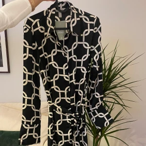 Svartvit långklänning med grafiskt mönster - Snygg långklänning från H&M i svart med vitt grafiskt kedjemönster. Klänningen har långa ärmar, krage och knytband i midjan för en markerad siluett. Tillverkad i återvunnen polyester som ger ett mjukt och följsamt fall.