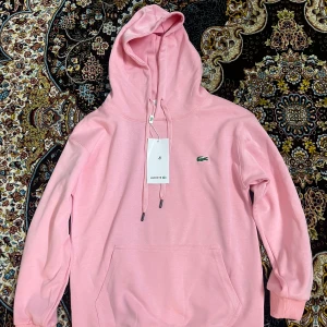 Rosa Lacoste - Säljer en snygg rosa hoodie från Lacoste med klassisk krokodillogga på bröstet. Tröjan har huva med snörning, känguruficka och ribbade muddar. Perfekt för dig som vill ha en stilren och bekväm look. Den är lite i storleken, den är xs skulle jag säga och är lite kort vid armarna men perfekt vid midjan för en S person. Jag är 175 och väger 65kg och den är jätte lite på mig. Den är oanvänd