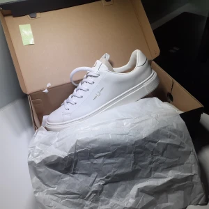 Vita sneakers från Fred Perry - Stilrena vita sneakers från Fred Perry med klassisk design. Skorna har snörning, rund tå och är tillverkade i slätt skinn. Fred Perry-loggan syns i guld på sidan och på sulan. Perfekta för en clean och fräsch look. PRISET ÄR EJ HUGGET I STEN