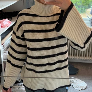 Randig polotröja från Ellos - Säljer en ribbstickad polotröja från Ellos i beige med svarta ränder. Tröjan har breda ärmar, hög polokrage och en liten slits i sidan. 