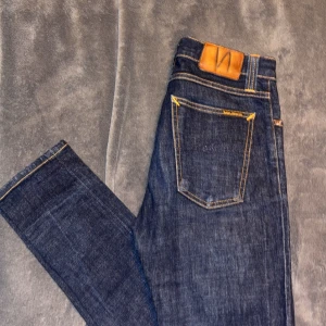 Nudie Jeans W28 L30 - Säljer ett par mörkblå jeans från Nudie Jeans i modellen W28 L30. Jeansen har klassisk femficksdesign, kontrastsömmar och läderpatch bak. Tillverkade i 100% ekologisk bomull för en schysst känsla och look.