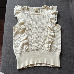 Vit stickad ärmlös topp Holly & Whyte - Supersöt vit stickad topp från Holly & Whyte med volangdetaljer på axlarna och ett snyggt hålmönster över hela plagget. Vid halsen har den lite smink fläckar men verkligen inget man tänker på. Perfekt till vardags eller om man vill klä upp sig!!