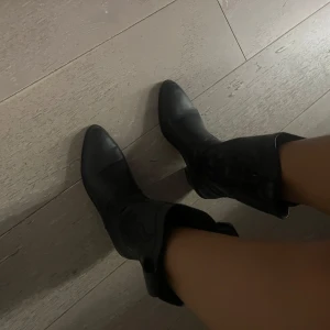 Svarta cowboy boots med klack - Säljer ett par svarta cowboy boots i skinnimitation med snygga broderade detaljer på skaftet. Bootsens spetsiga tå och stabila klack ger en cool western-vibe. Perfekta att styla till jeans eller klänning för en trendig look.