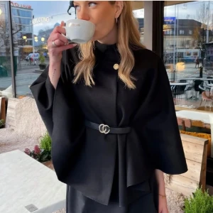 Poncho med bälte och guldknapp - Snygg poncho från Missäy med hög krage, guldknapp vid halsen och knytskärp i midjan i svart. Jackan har vida ärmar och en stilren, elegant look som lyfter vilken outfit som helst.