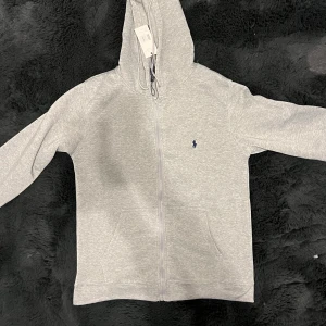 Grå hoodie från Polo Ralph Lauren - Snygg ljusgrå hoodie från Polo Ralph Lauren med dragkedja och klassisk blå logga broderad på bröstet. Tröjan har luva med snörning, känguruficka och långa ärmar. Perfekt för chill dagar och enkel att matcha med jeans eller joggers.
