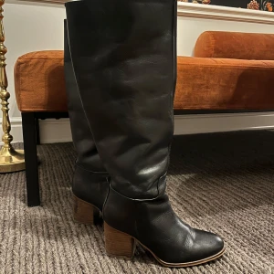 Svarta höga stövlar med klack - Snygga svarta stövlar i skinn med rund tå och bred träfärgad klack. Stövlarna har en klassisk och stilren look med lite slouchig passform på skaftet. Perfekta att styla med kjol eller jeans för en trendig vibe.