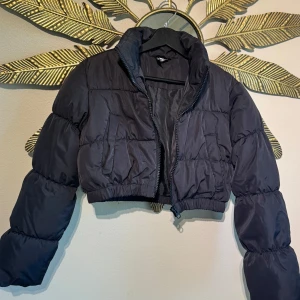 Svart croppad pufferjacka - H&M Divided  - Oanvänd. Snygg svart croppad pufferjacka med hög krage och dragkedja framtill. Jackan har långa puffiga ärmar och elastisk midja för en trendig siluett. Perfekt för dig som vill ha en cool och modern look under kyliga dagar.