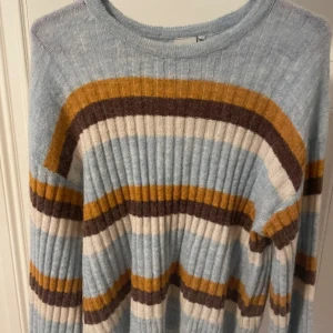 Randig ribbstickad tröja i pastell - Superfin ribbstickad tröja med breda ränder i ljusblått, vitt, brunt och senapsgult. Tröjan har rund halsringning och långa ärmar. Perfekt för dig som gillar retrovibbar och mjuka material. Använd 2-3 gånger 