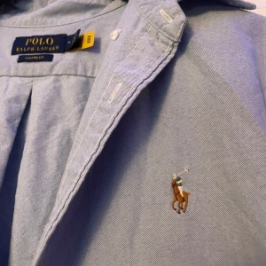 Blå kortärmad skjorta Polo Ralph Lauren - Snygg ljusblå kortärmad skjorta från Polo Ralph Lauren med klassisk krage och broderad logga på bröstet. Skjortan har custom fit-passform och knäppning framtill. Perfekt för en clean och stilren look. Nästan oanvänd 