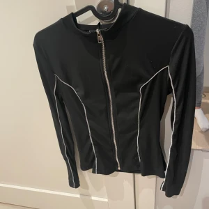Svart ziptröja med vita sömmar - Svart långärmad ziptröja med vita kontrastsömmar och hel dragkedja framtill. Tröjan har en smal passform och ribbad struktur, perfekt för en sportig och stilren look.