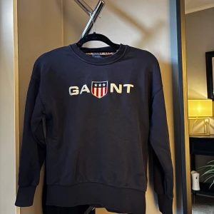 Mörkblå sweatshirt från Gant - Säljer en mörkblå sweatshirt från Gant med broderad logga och sköld på bröstet. Tröjan har rund halsringning, långa ärmar och ribbade muddar vid ärmslut och nederkant. Perfekt för en avslappnad och stilren look.
