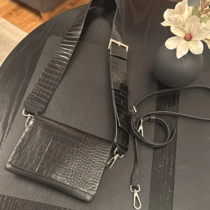 Svart croco axelväska med bred rem+smal rem Carin Wester - Snygg svart axelväska i croco-mönstrat skinn med bred justerbar rem och silverfärgade detaljer från Carin Wester. Väskan har även en smalare avtagbar rem och dragkedja på insidan för extra säkerhet. Perfekt för dig som vill ha en stilren och trendig accessoar.