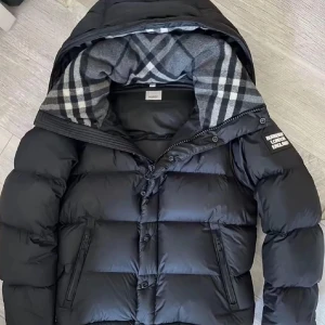 Svart pufferjacka från Burberry - Säljer en svart pufferjacka från Burberry med klassiskt rutigt foder i huvan och tydlig logga på ärmen. Jackan har dragkedja, knappar och två snedställda fickor framtill. Perfekt för kalla dagar och riktigt snygg streetstil. Man kan ta av armarna på jackan och användas som en jätte snygg väst. Skicka DM för bild på kvittot.