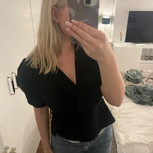 Svart med puffärm - Snygg svart med korta puffärmar och markerad midja genom knyt. Blusen har en djup v-ringning och en stilren, elegant look. Perfekt att styla med jeans eller kjol för en trendig outfit.