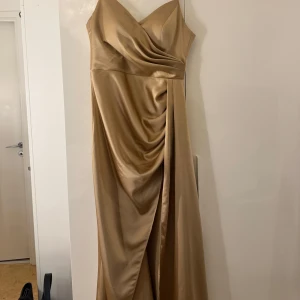 Champagnefärgad långklänning med slits - Elegant långklänning i champagnefärgad satin med smala axelband, v-ringning och draperad framsida. Klänningen har en snygg slits på ena sidan och figurnära passform som ger en lyxig känsla. Perfekt för bal eller speciella tillfällen. För fler bilder skriv privat! Kan gå ner i priset så skriv så kan det diskuteras.