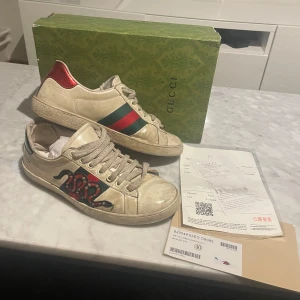 Gucci Ace sneakers med orm  - Gucci Ace sneakers i vitt skinn med klassiska gröna och röda ränder på sidan samt en broderad orm i rött och svart. Baksidan har metallicröd detalj. Skorna har snörning och platt sula. Kommer med originalkartong och kvitto. Passar även 42 och 43