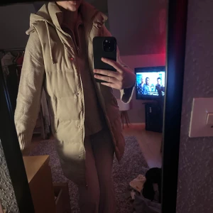 Beige lång pufferjacka med huva - Säljer en beige lång väst med huva och snörning. Jackan har dragkedja och knappar framtill, quiltad design. Perfekt för kyliga dagar och riktigt snygg till ett par jeans eller cargobyxor.